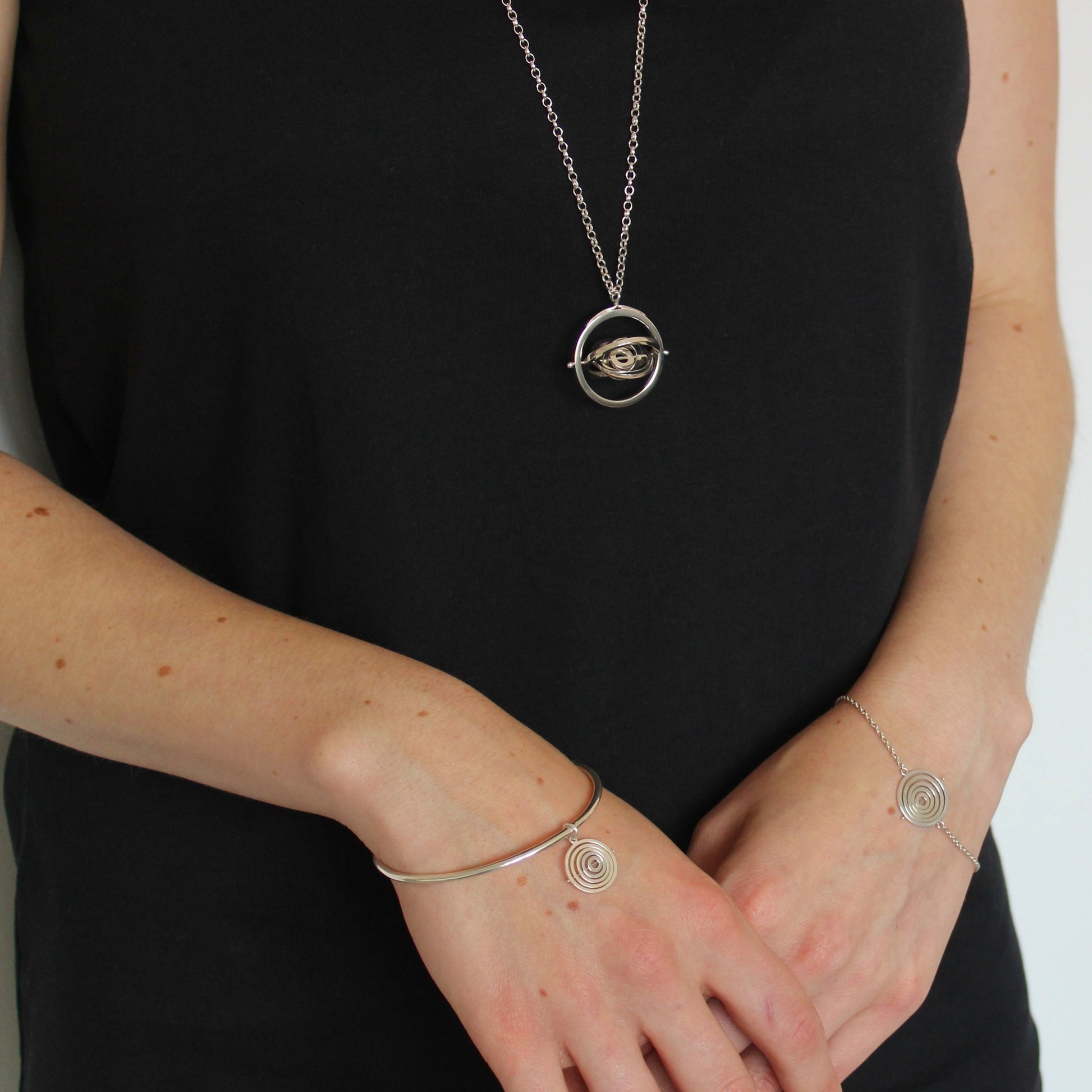 Model wearing hallmarked silver kinetic concentric circles pendant hanging below bust & matching charm bangle & bracelet